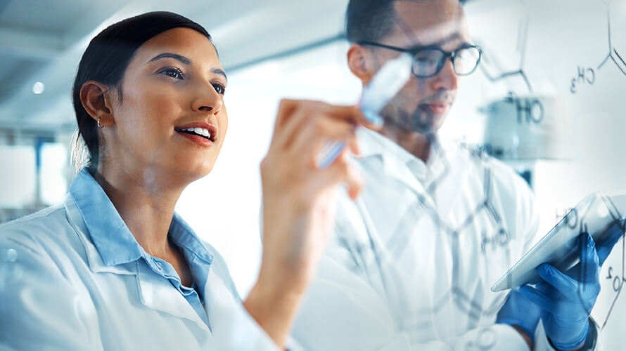 Two professionals in lab coats observe a computer screen, engaged in discussion in a lab environment.